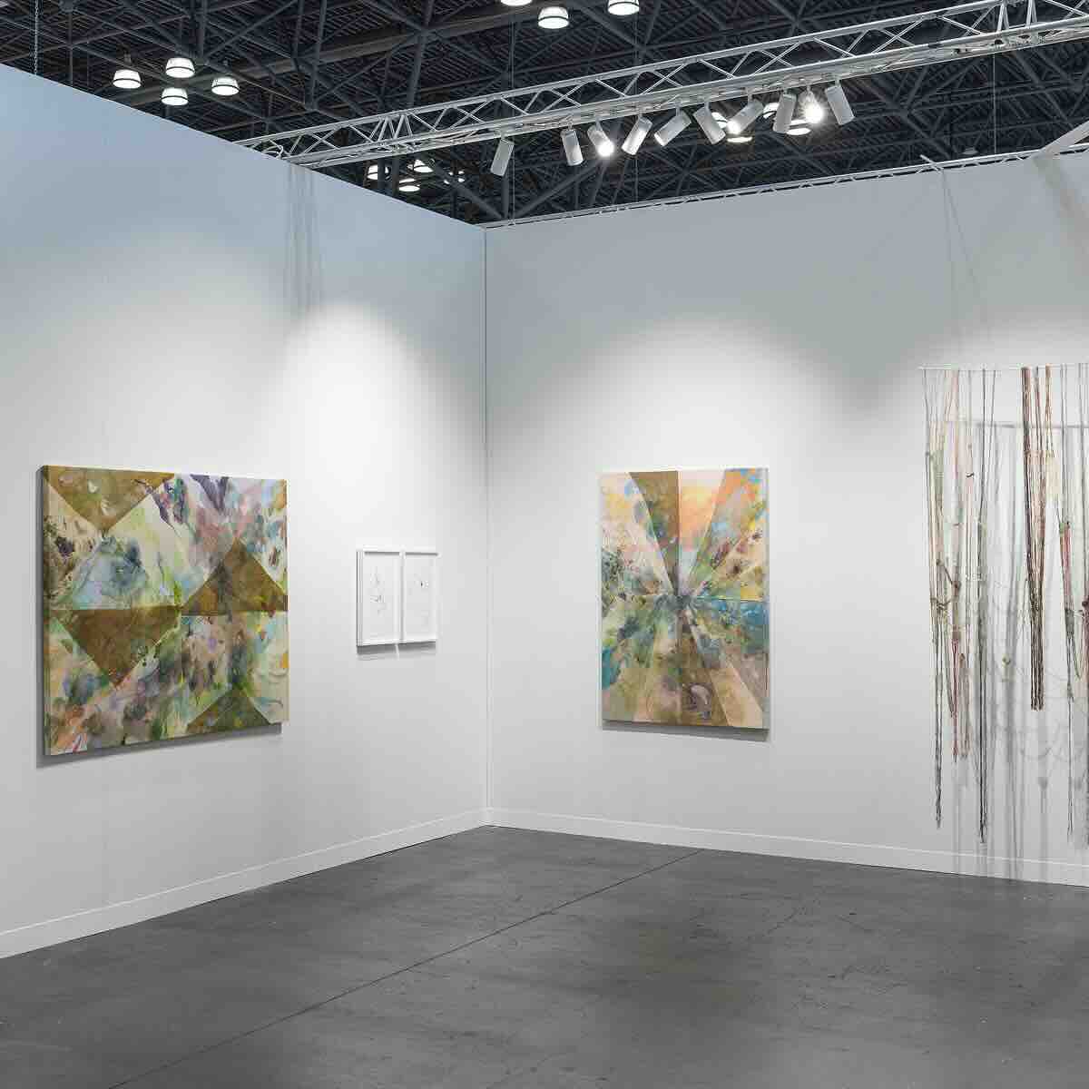 Gallery corner with three abstract mixed-media artworks on white walls: two colorful geometric paintings and one installation with hanging strings or threads.