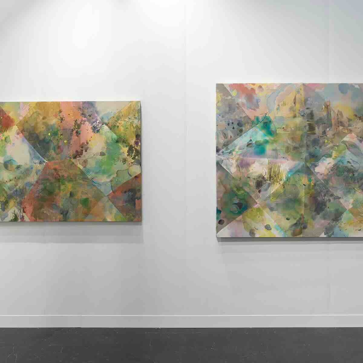 Two abstract paintings with geometric patterns and multicolored washes of pastel colors including greens, yellows, pinks, and blues hang on a white gallery wall above a dark floor.