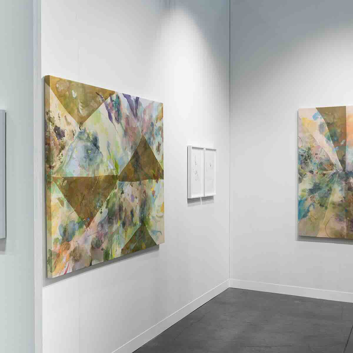 White gallery walls displaying abstract paintings with geometric shapes and muted colors like green, brown, and blue, illuminated by spotlights; a pair of small framed sketches or drawings hang between the larger paintings.