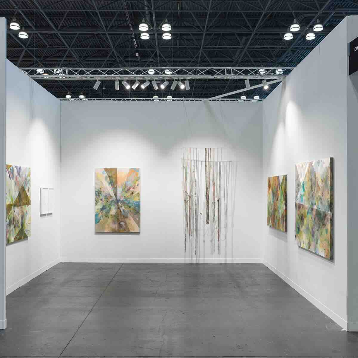 Contemporary art gallery corner with white walls and concrete floor, featuring abstract paintings with muted pastels and earthy tones on the side walls and a hanging sculptural installation of vertical strands in the center wall.