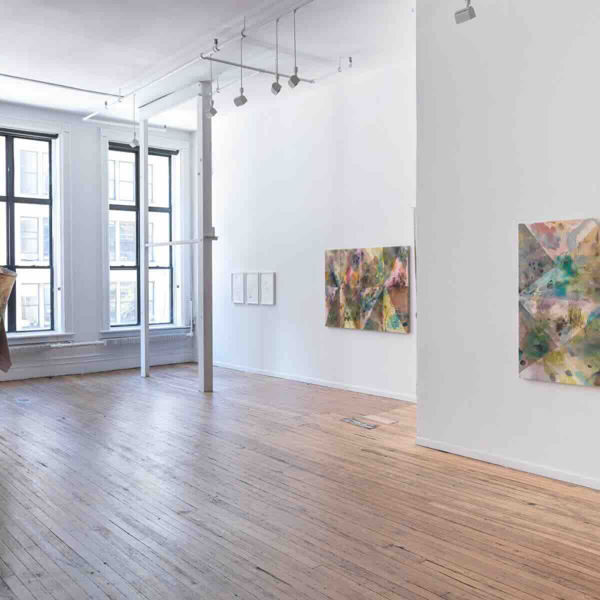 Bright, spacious art gallery with wooden floors, large windows, and abstract colorful paintings on white walls.