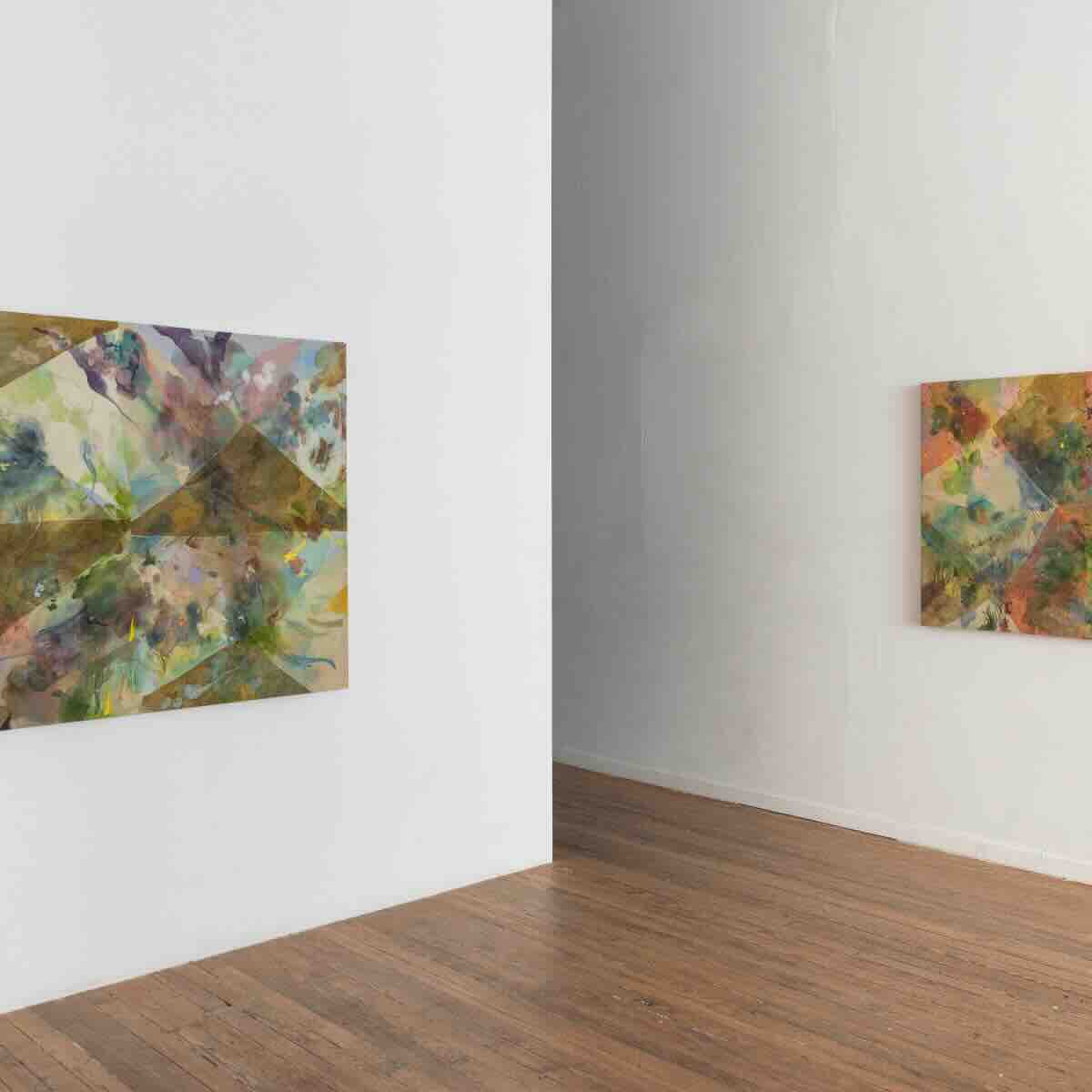 Interior view of a white gallery room with wooden flooring, featuring two abstract paintings with geometric shapes and colorful, textured patterns mounted on adjacent walls.
