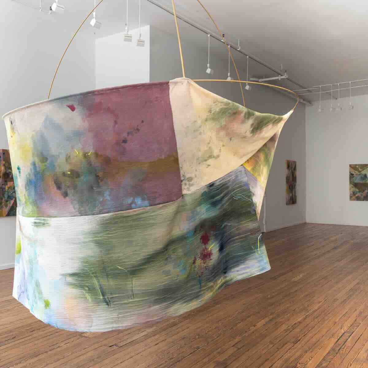 Large curved fabric piece with abstract watercolor-like patterns in pastel colors suspended by thin rods in a white gallery space with wooden floors and additional framed artworks on the walls.