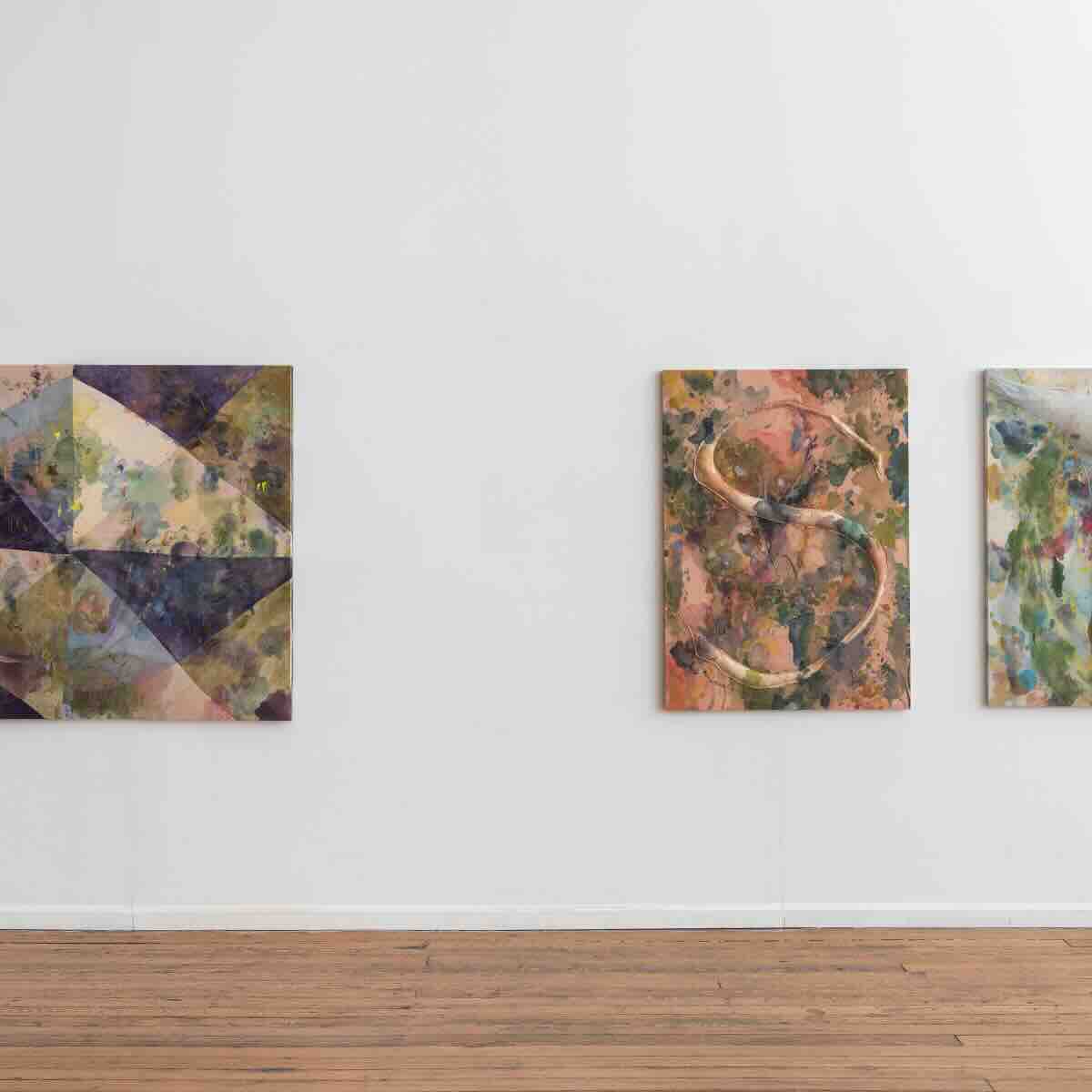 Three abstract paintings hang on a white gallery wall above a wooden floor; the left painting features geometric shapes in muted colors, the middle one displays organic curved forms on a textured brown and green background, and the right one shows a mix of blue, green, and orange hues with a textured appearance.