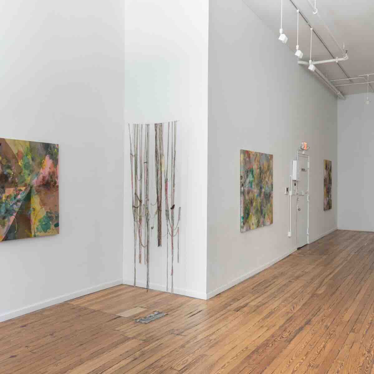 Minimalist art gallery interior with wooden floors and white walls featuring three abstract paintings and a hanging string installation in a corner.