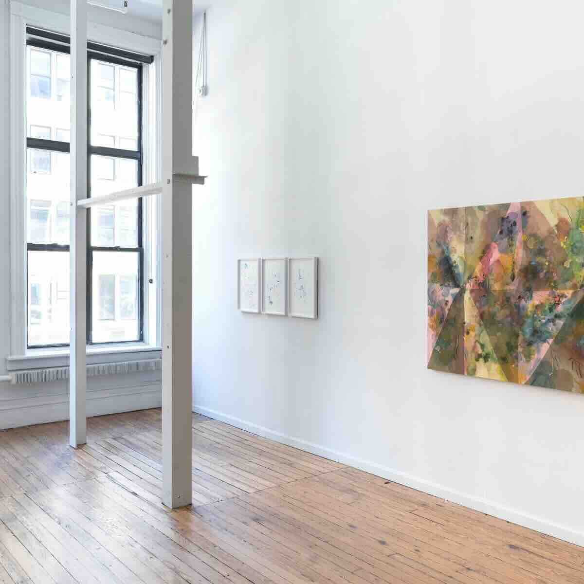Bright art gallery room with large window, wooden floor, white walls, three small framed abstract drawings on one wall, and a large colorful abstract painting on another wall.