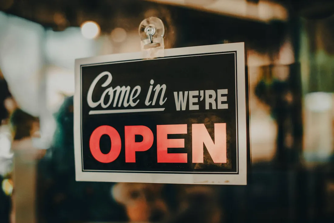 Sign on glass door reading 'Come in we're OPEN' with blurred warm lights in background.