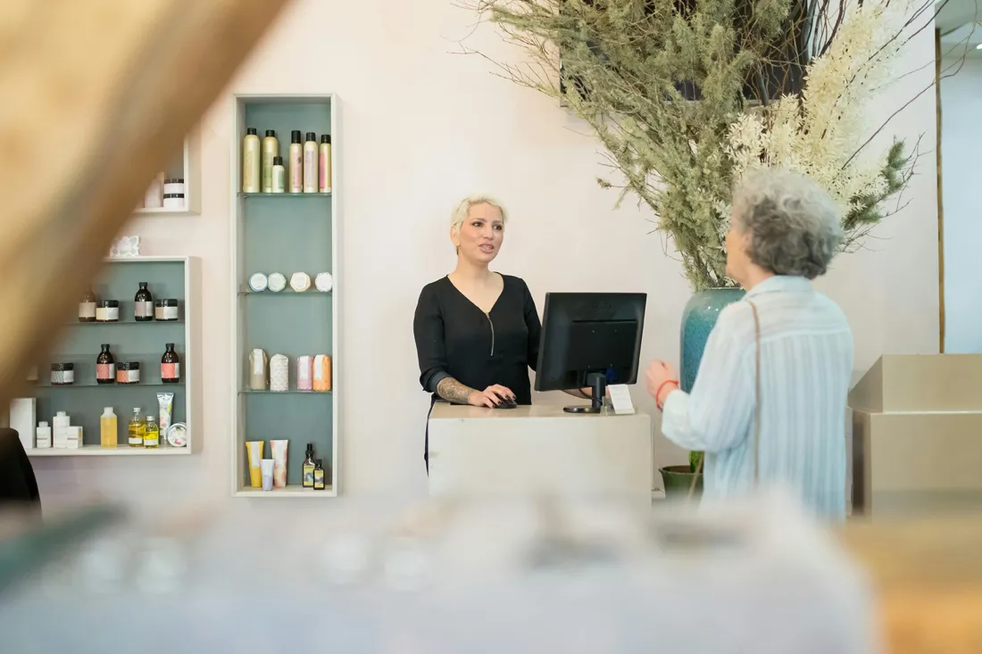 Woman with tattoos at a store counter talking to an older woman near shelves with skincare products.