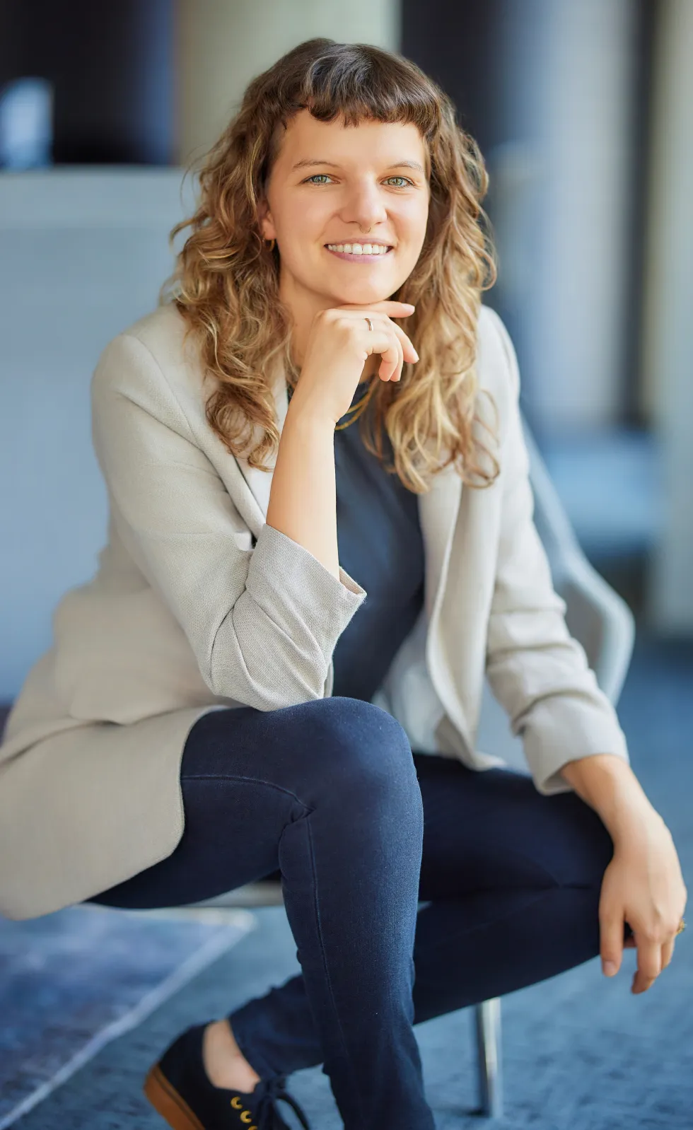Smiling woman with curly hair sitting cross-legged on a chair wearing a beige blazer and dark pants.