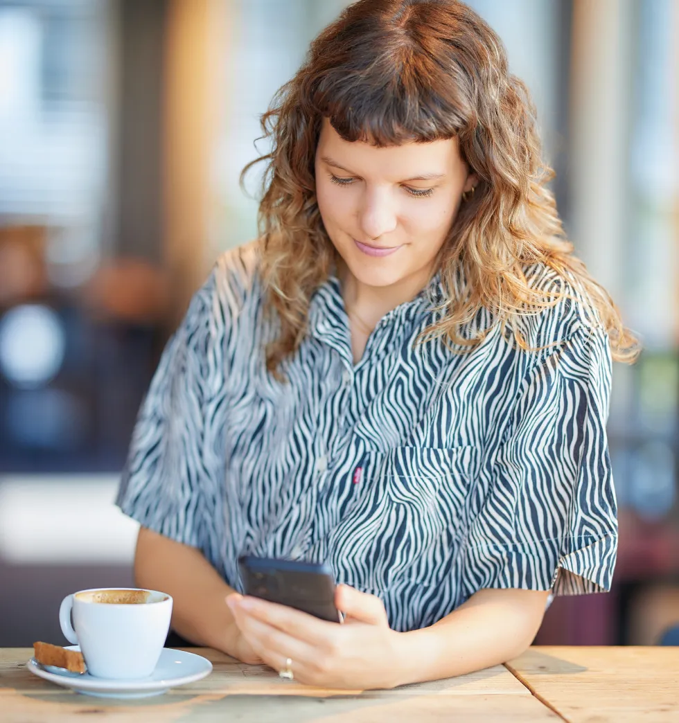 Woman with curly hair wearing a zebra-striped shirt looking at her smartphone with a cup of coffee on the table.