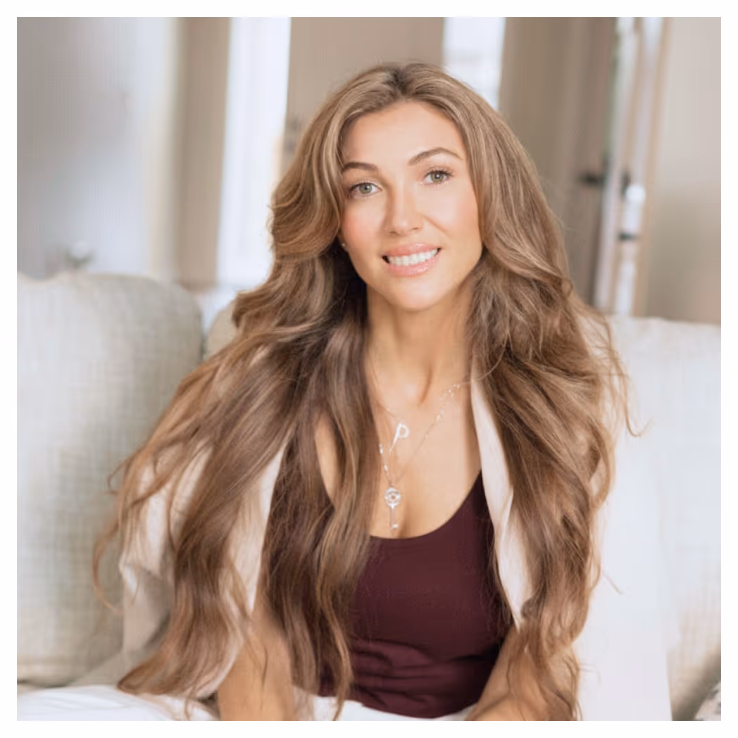 Woman with long wavy hair wearing a maroon tank top and light-colored open jacket, smiling while seated on a couch.