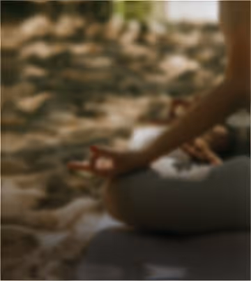 Person sitting cross-legged on a mat outdoors, meditating with hands resting on knees in a mudra gesture.