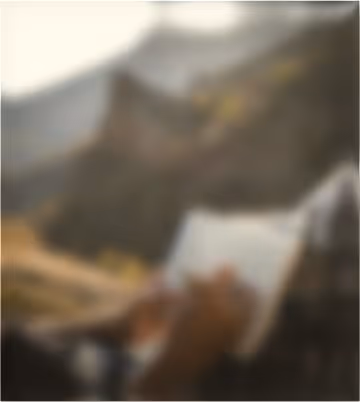 Blurred image of a person holding an open book with a mountainous landscape in the background.