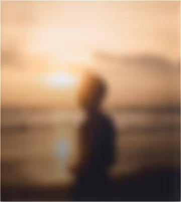 Blurry silhouette of a person standing by the water during sunset with an orange sky.