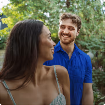 Smiling young man in a blue shirt looking at a woman with long dark hair outdoors surrounded by green trees.