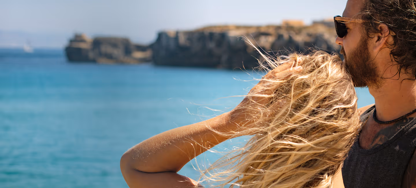 Couple standing close by the ocean with cliffs in the background, the man wearing sunglasses and the woman with windblown blonde hair.