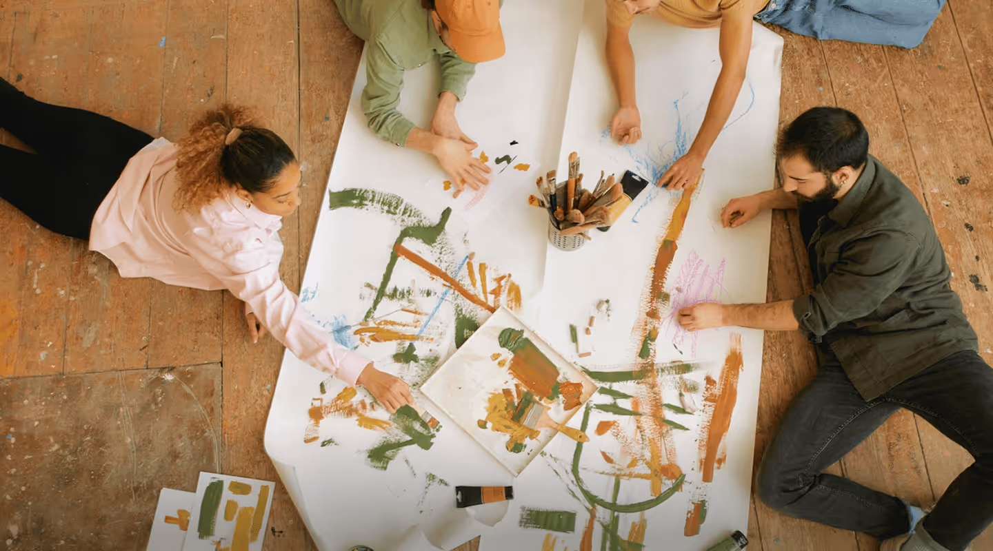 Four people lying on a wooden floor collaboratively painting on a large white sheet with brushes and colorful paint strokes.