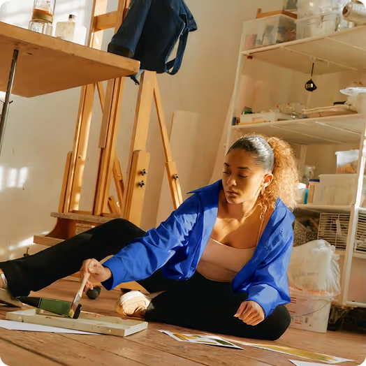 Woman in a blue jacket sitting on the floor painting on a canvas in a well-lit art studio.