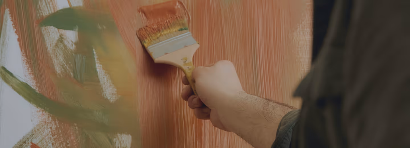 Hand holding a paintbrush applying broad strokes of reddish-brown paint on a surface with mixed green and white paint areas.