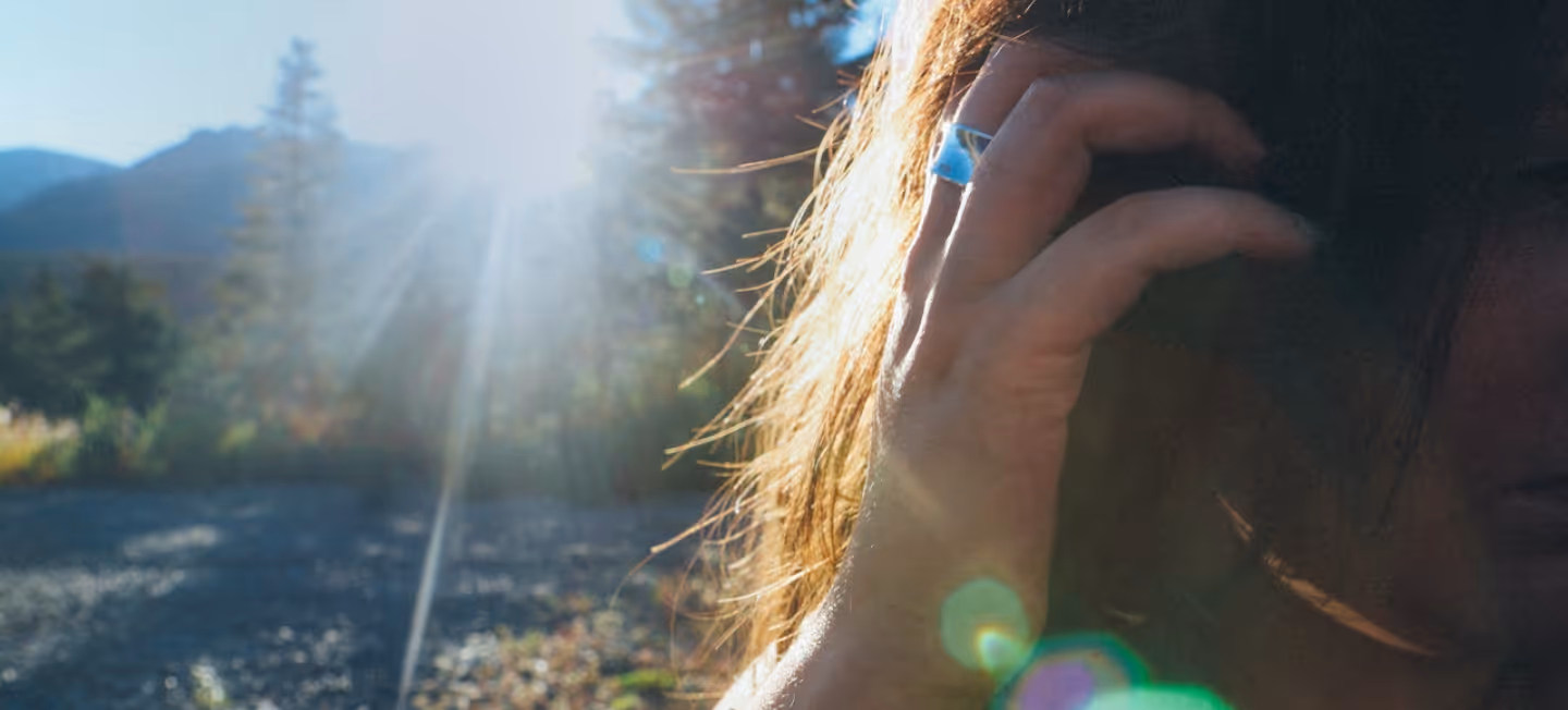 Sun shining over a forested mountain landscape with a person partially visible touching their hair, with lens flare effects.