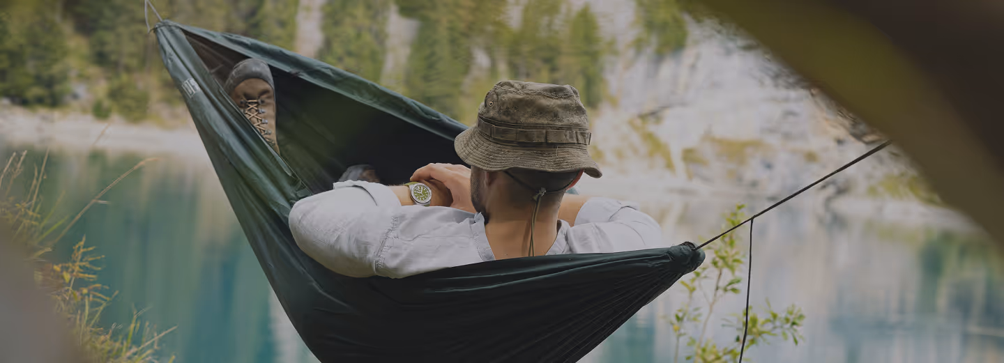 Man wearing a hat and boots relaxing in a hammock by a calm lake surrounded by trees and cliffs.