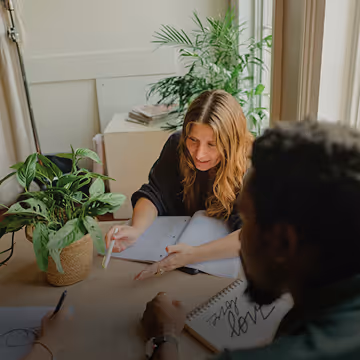 Two people sitting at a table with notebooks, discussing or working together near green plants in a well-lit room.