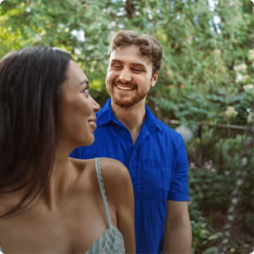 Smiling man in a blue shirt looking at a woman in a patterned dress outdoors with green foliage in the background.