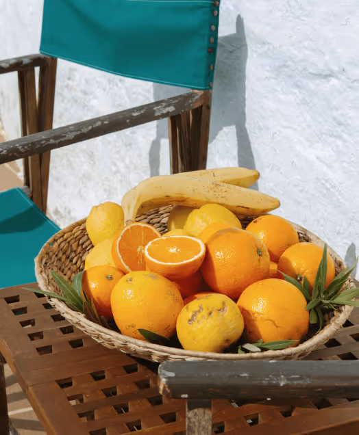 Wicker basket filled with oranges, lemons, and bananas on a wooden table with a green chair in the background.