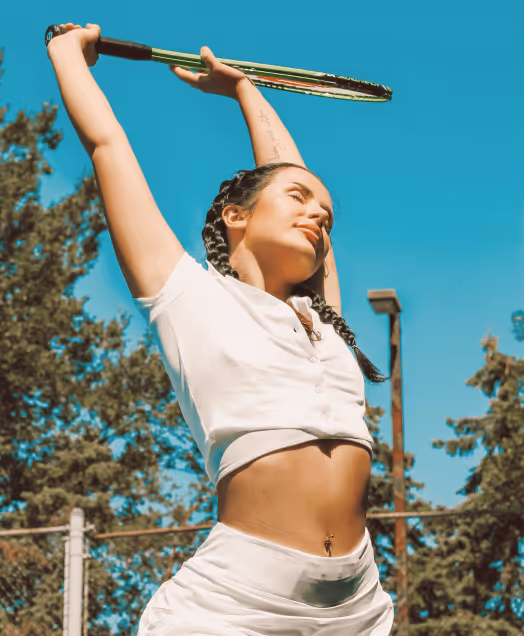 Woman with braided hair stretching arms above head while holding a tennis racket outdoors under clear blue sky.