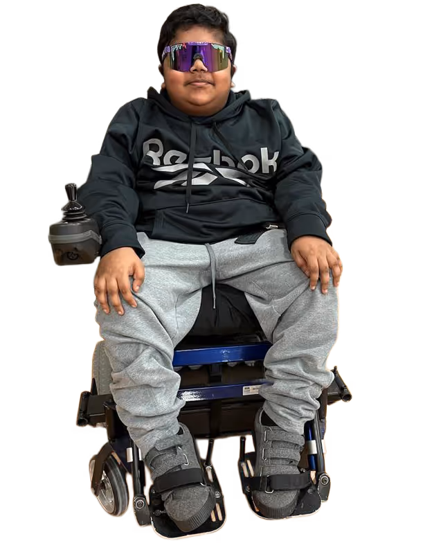 Boy wearing a black Reebok hoodie, gray sweatpants, and colorful reflective sunglasses sitting in a motorized wheelchair.