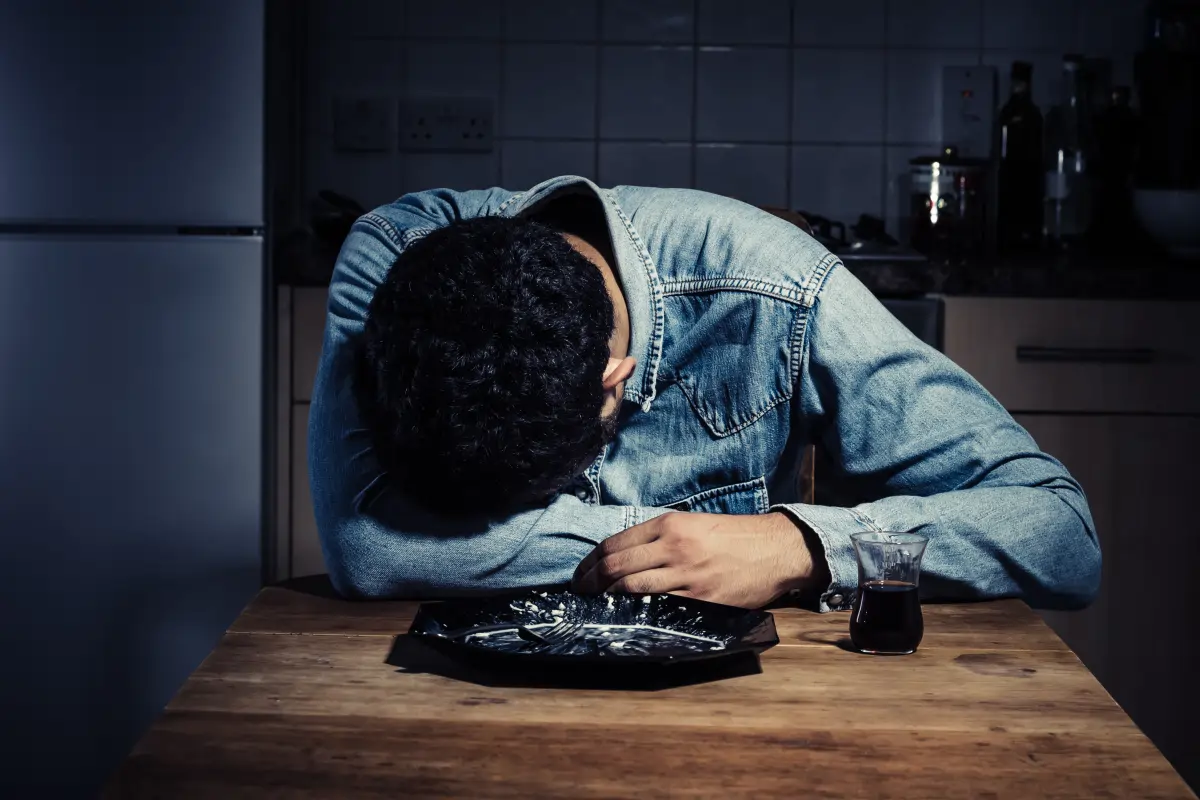 Man slumped over table with drink in dim kitchen, representing stress-related erectile dysfunction, substance use, and common psychological causes of ED in men.