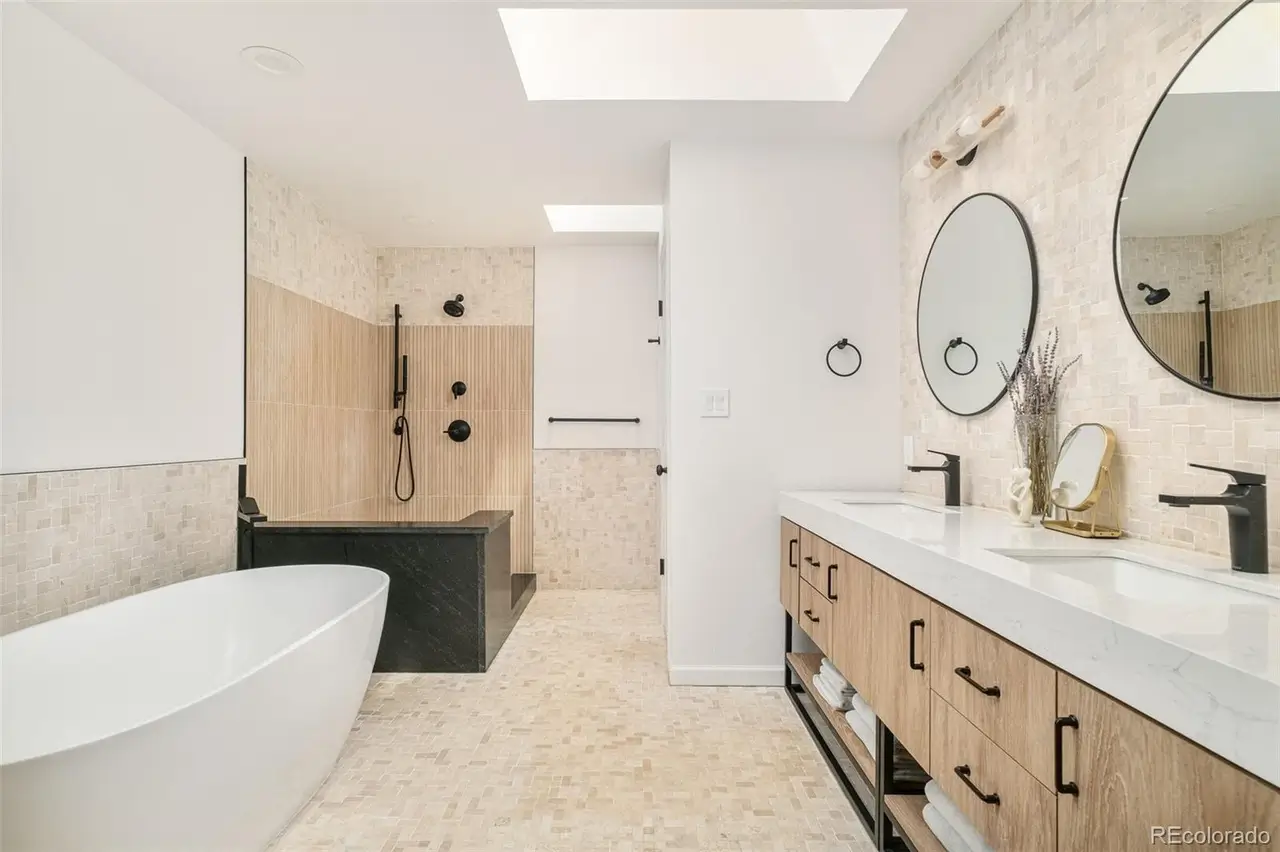 Contemporary bathroom with freestanding white soaking tub, walk-in shower with black fixtures, dual vanity with wood cabinetry and vessel sinks, round mirrors, and neutral tile walls and floors.