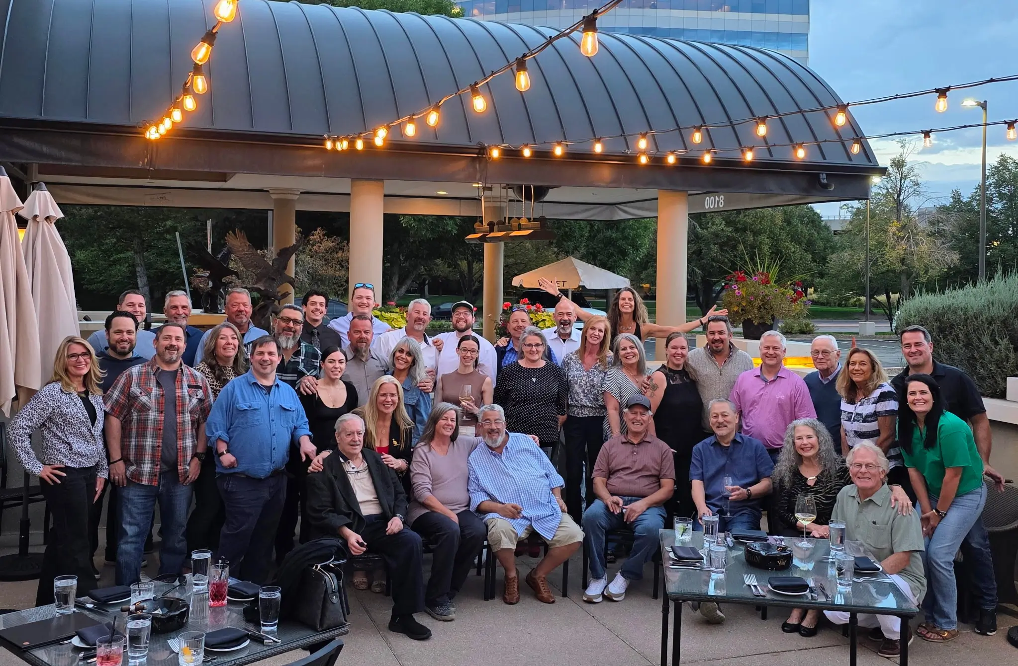 Group photo of Denver Dream Builders’ friends and family gathered outdoors at a patio restaurant, smiling together under string lights during an evening celebration.