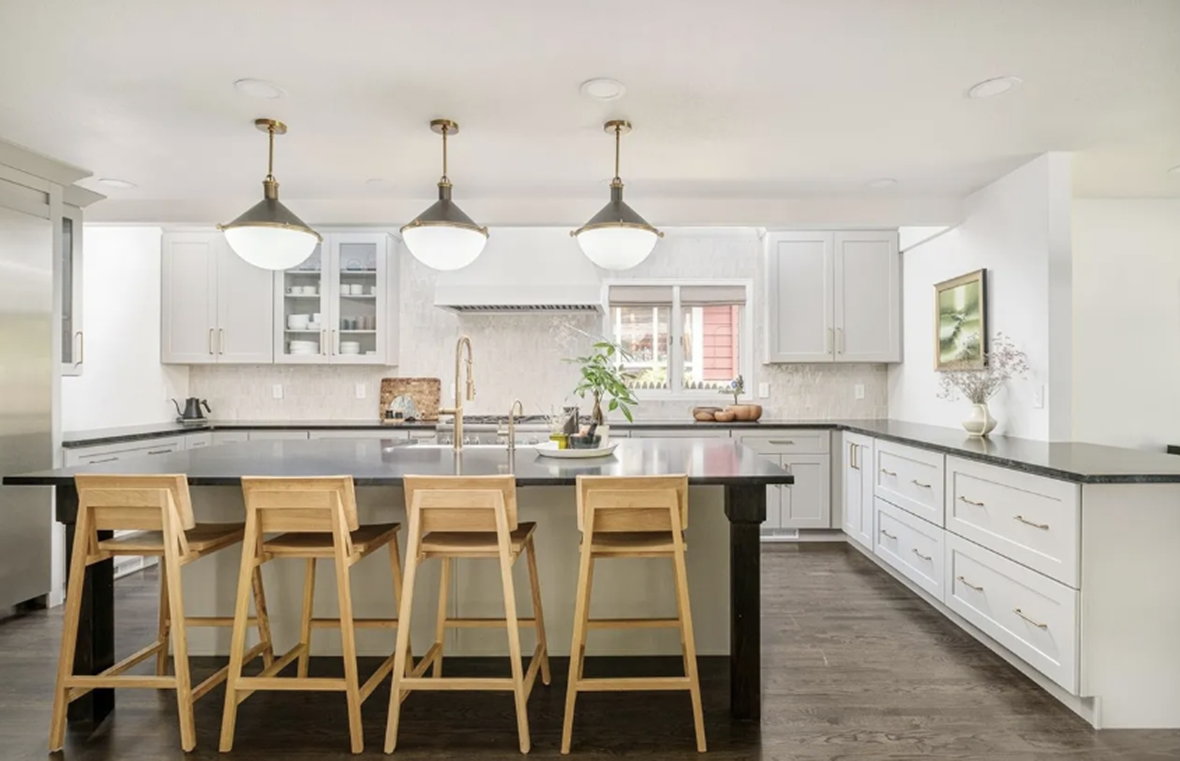 Modern kitchen with white shaker cabinets, dark stone countertops, farmhouse sink, stainless steel appliances, gas range with hood, pendant lights, and hardwood floors.