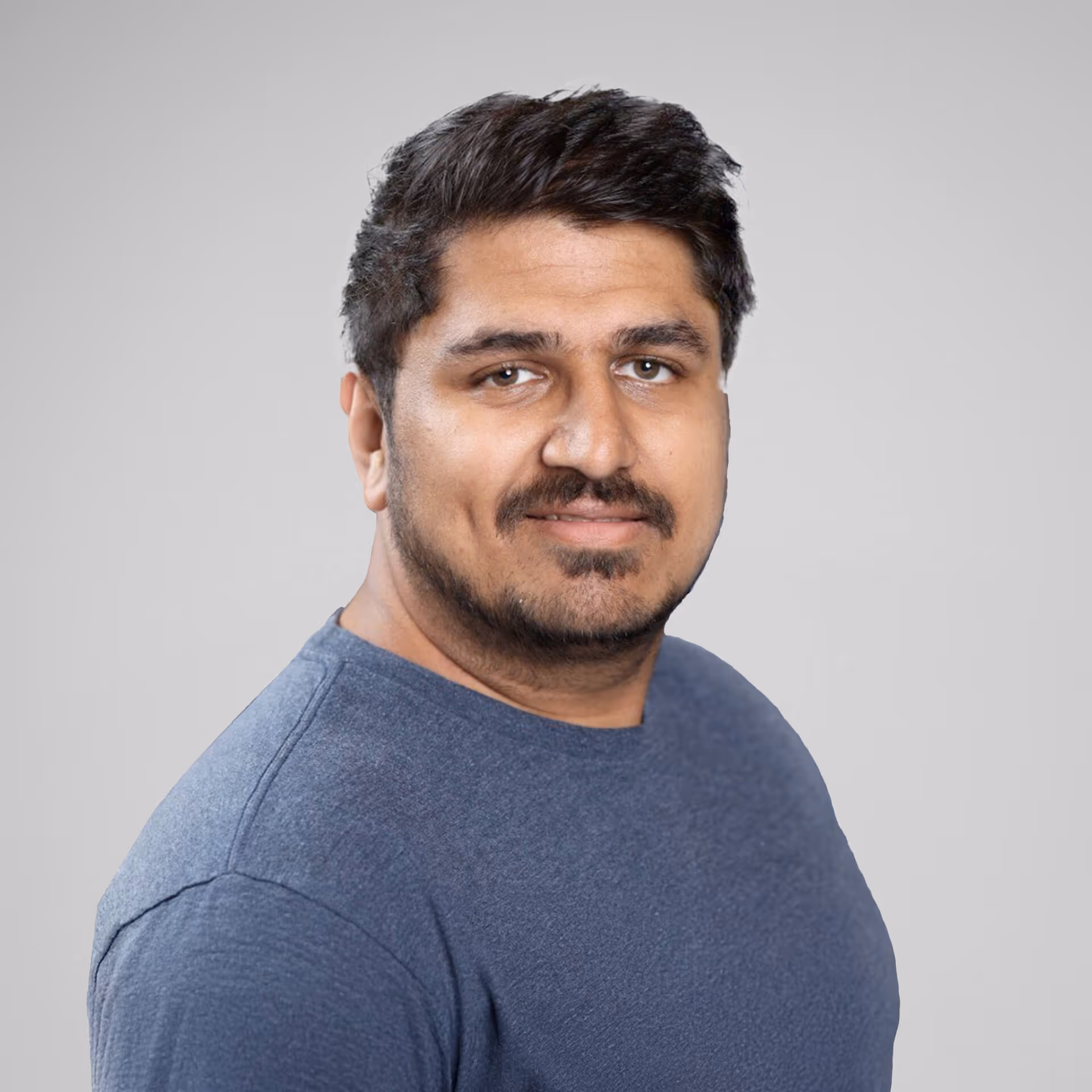 Portrait of a man with short dark hair and a beard wearing a blue shirt, against a plain light gray background.