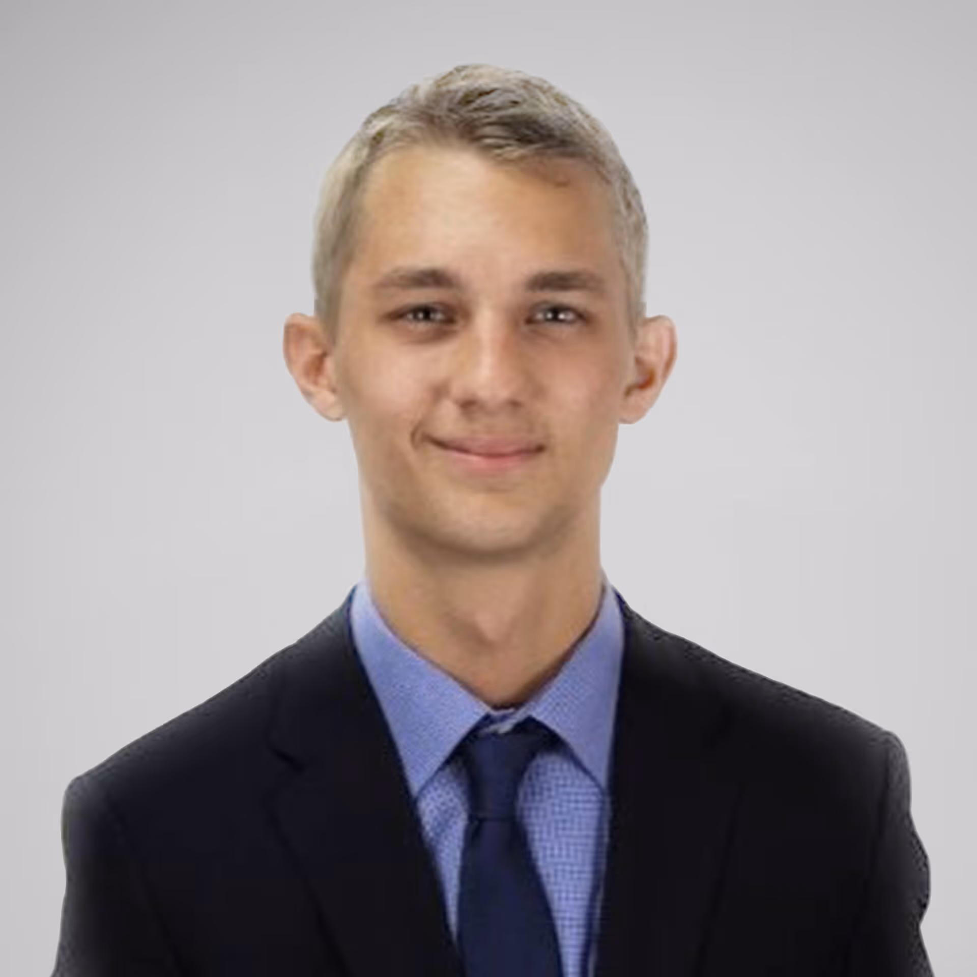 Young man with short blond hair wearing a black suit, blue shirt, and navy tie against a light gray background.