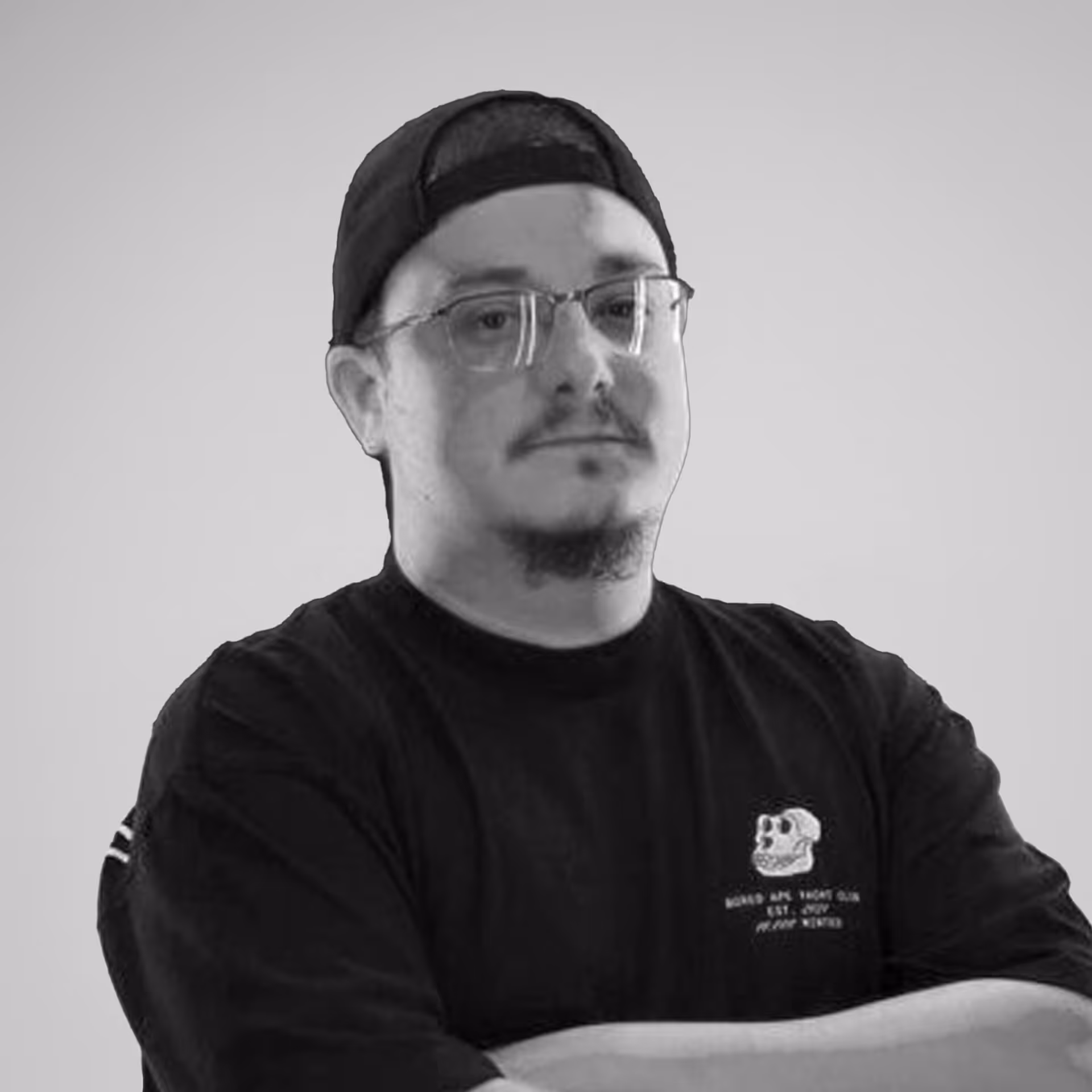 Man with glasses and a goatee wearing a backward cap and a black shirt with a small skull graphic and text, arms crossed.