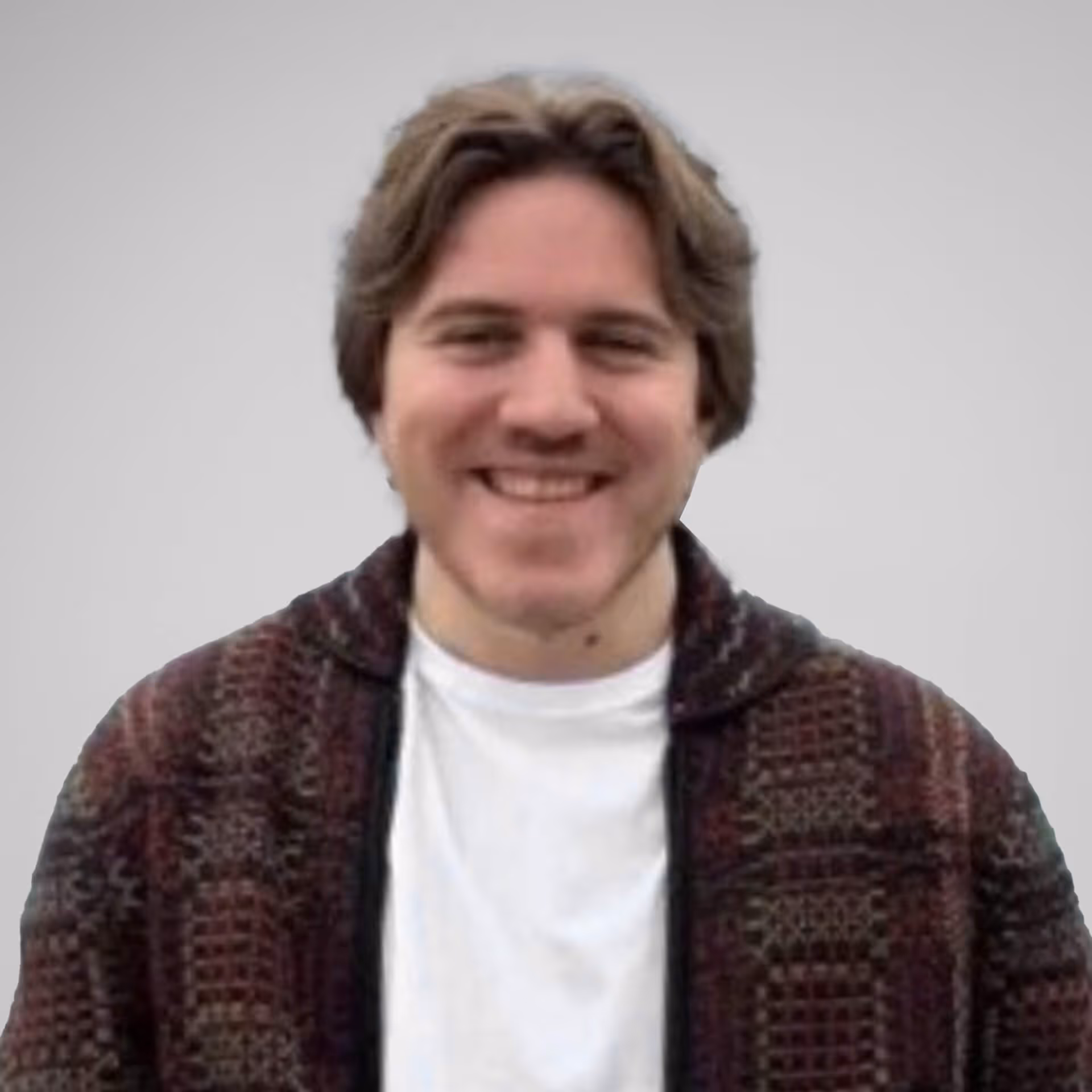 Smiling man with light brown hair wearing a patterned jacket over a white shirt in front of a gray background.