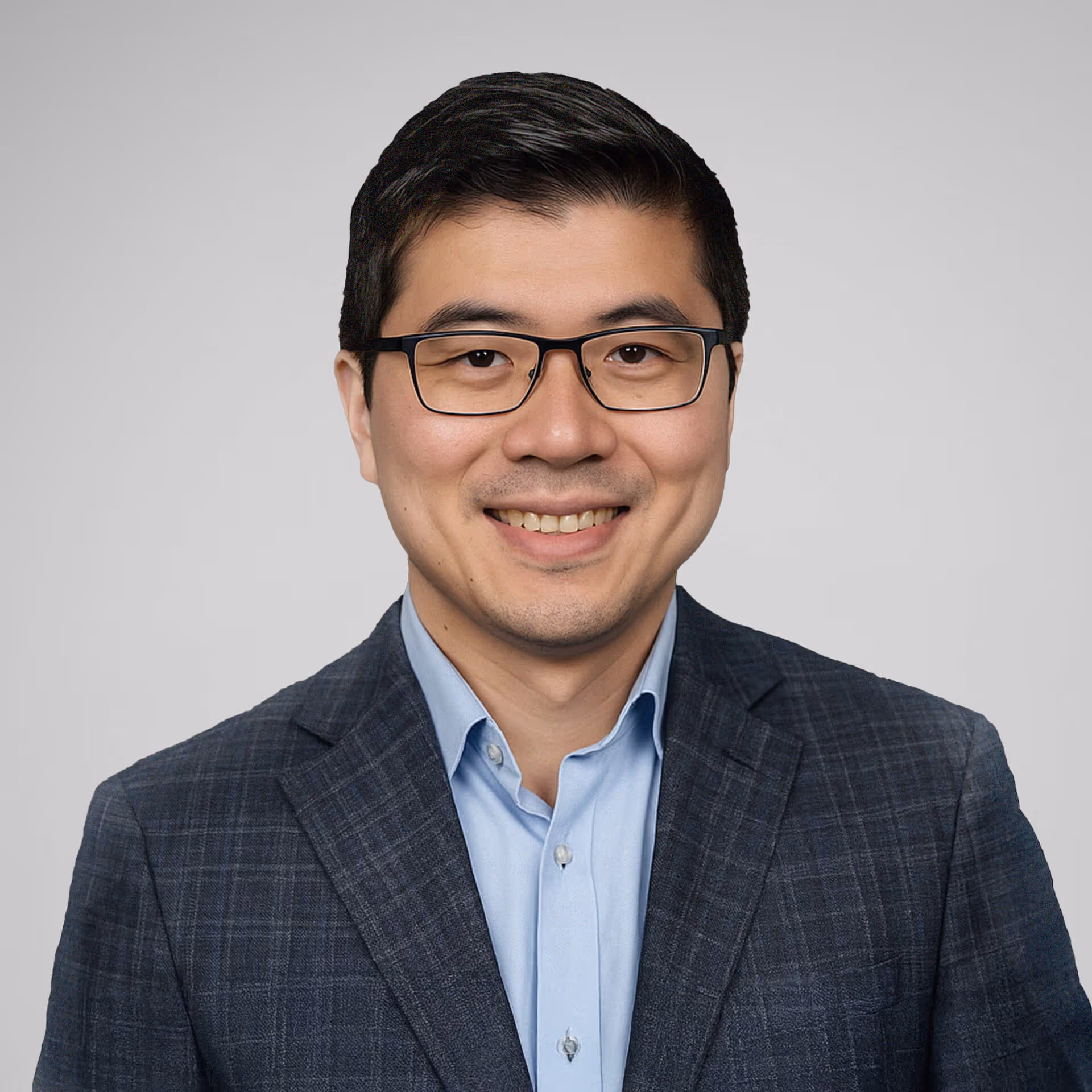 Smiling man wearing glasses, a light blue shirt, and a dark checked blazer, against a plain gray background.