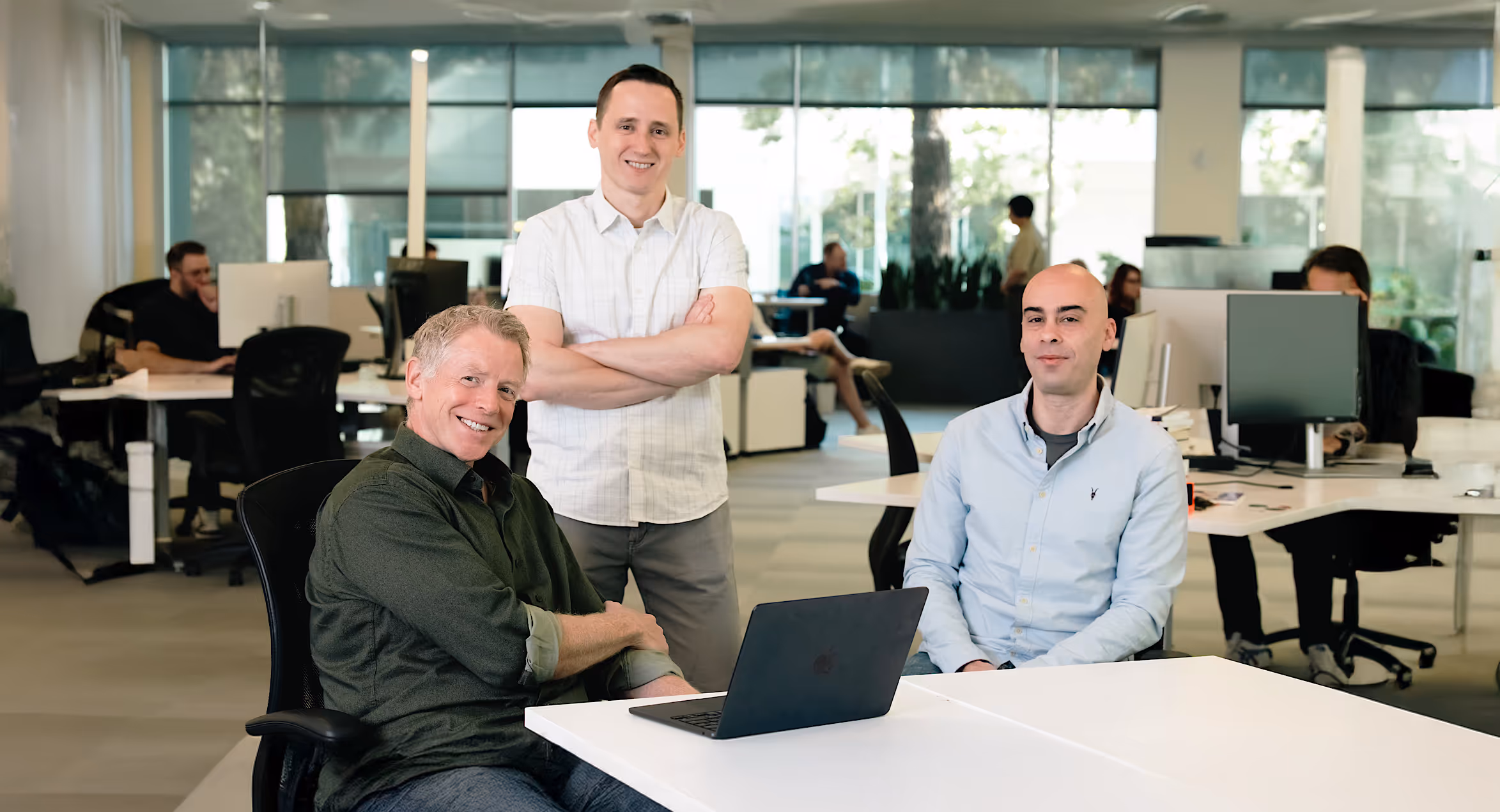 Three men in a modern office, two seated at a table with a laptop and one standing behind them with arms crossed.