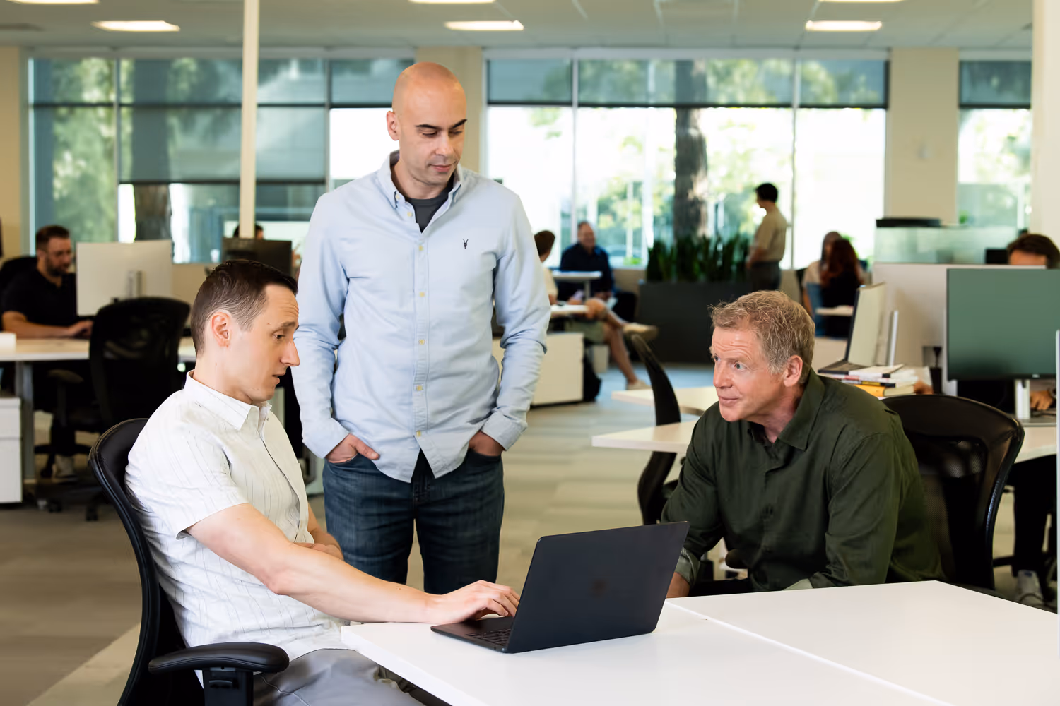 Three men in an office collaborating over a laptop, with one seated typing, one standing, and one seated leaning forward.