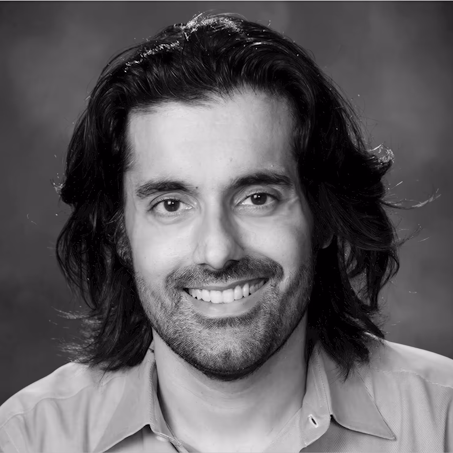 Black and white headshot of a smiling man with medium-length dark hair and a trimmed beard, wearing a collared shirt.