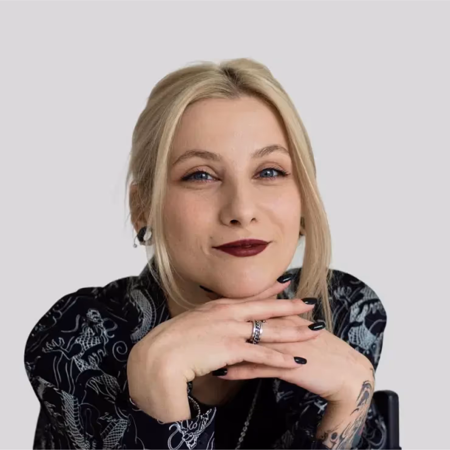 Smiling blonde woman with dark lipstick resting her chin on her hands, wearing a black patterned shirt and a tattoo on her forearm.