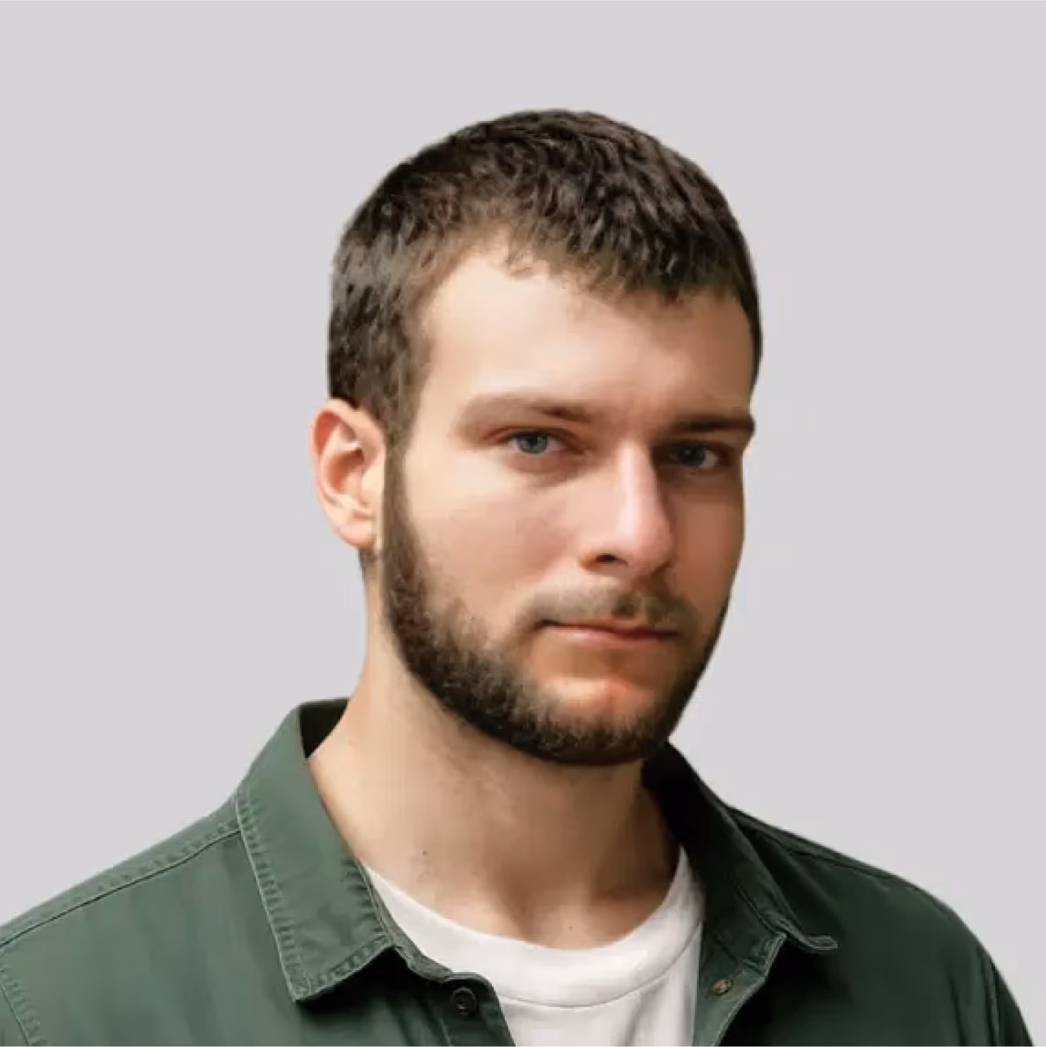 Young man with short brown hair and beard wearing a green jacket over a white shirt against a gray background.