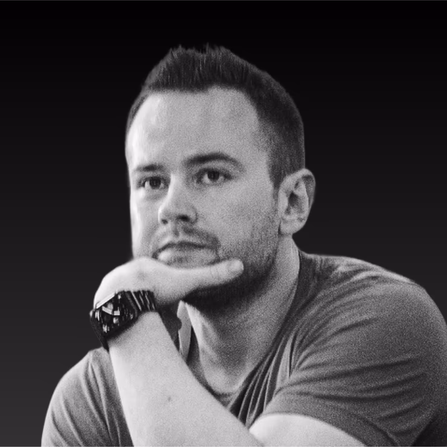 Black and white portrait of a man resting his chin on his hand, wearing a watch and a t-shirt, against a dark gradient background.
