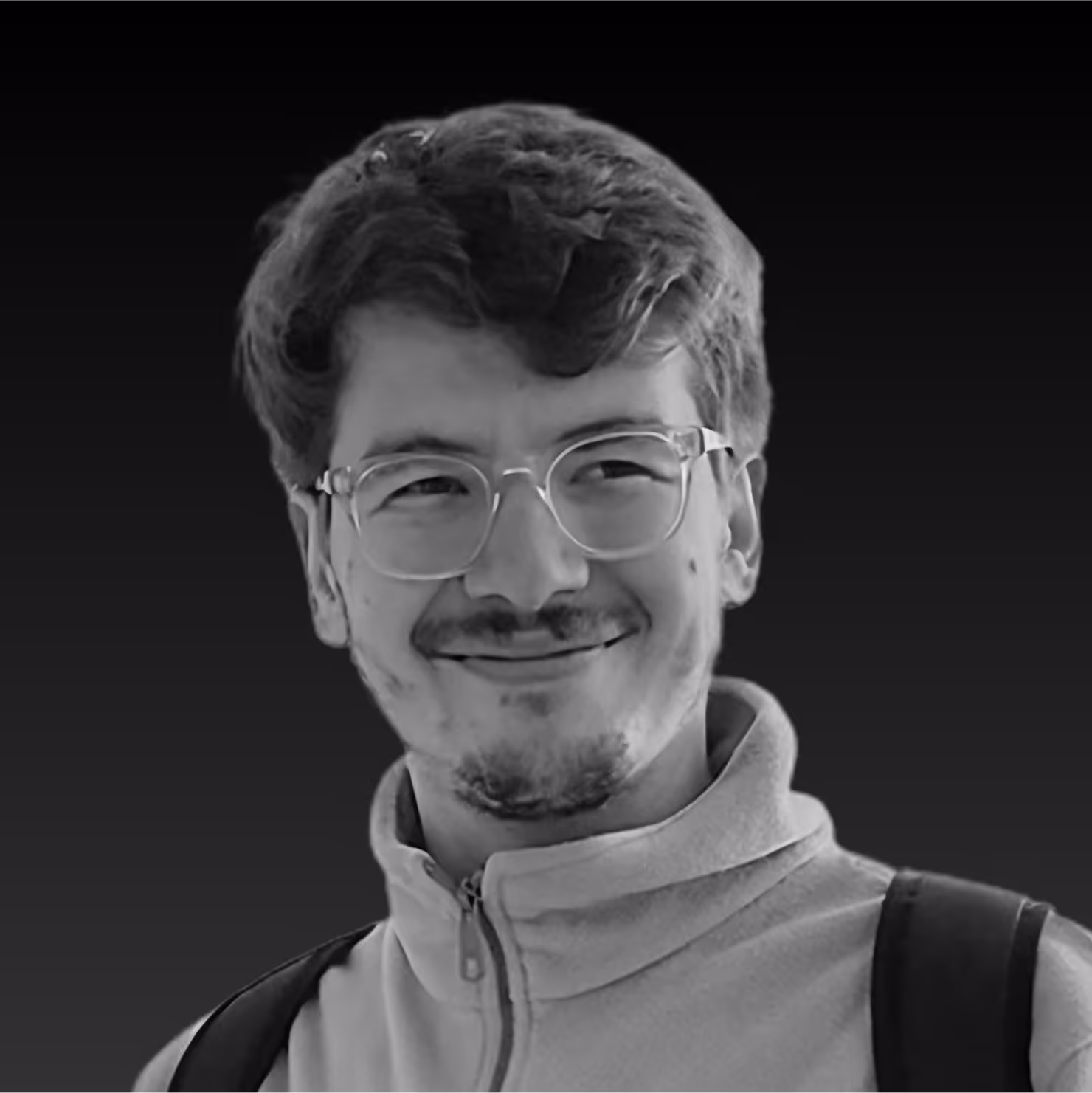 Smiling young man with glasses and facial hair wearing a zipped jacket and backpack against a dark background.