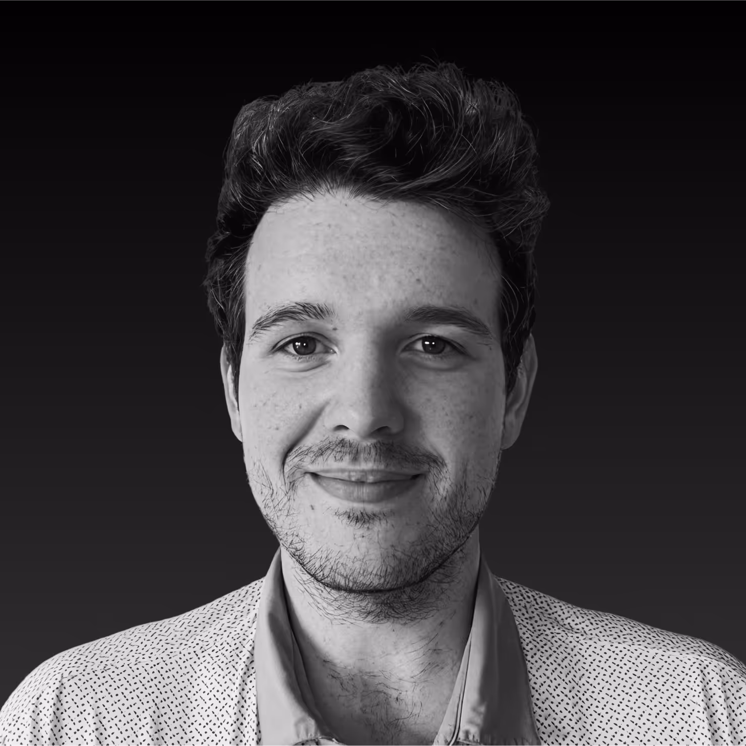 Close-up black and white portrait of a smiling young man with textured hair and light stubble, wearing a patterned shirt.