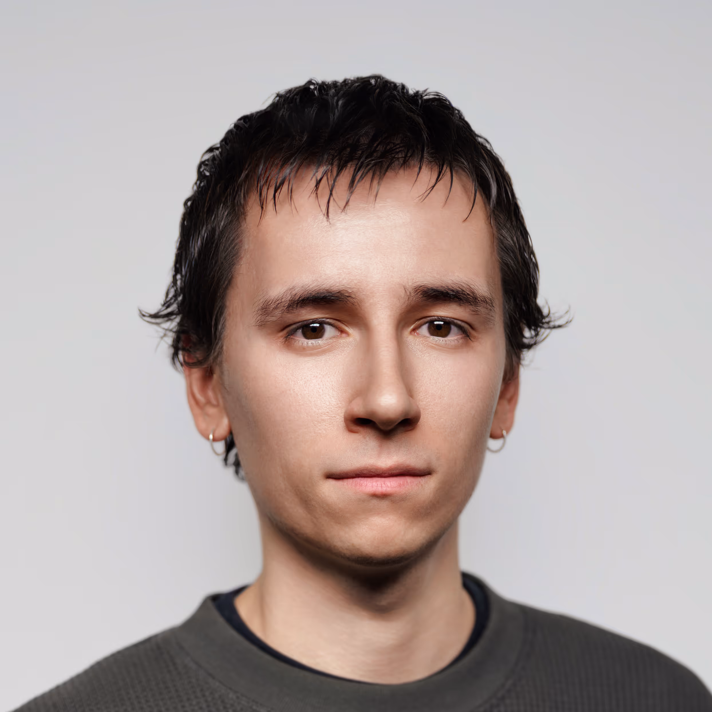 Close-up portrait of a young man with short dark hair, wearing small hoop earrings and a dark green sweater, against a plain light gray background.
