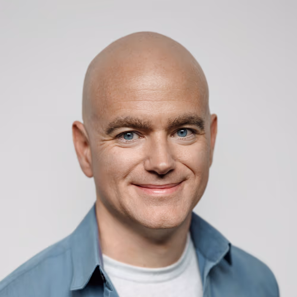 Smiling bald man with blue eyes wearing a light blue shirt over a white t-shirt against a plain background.