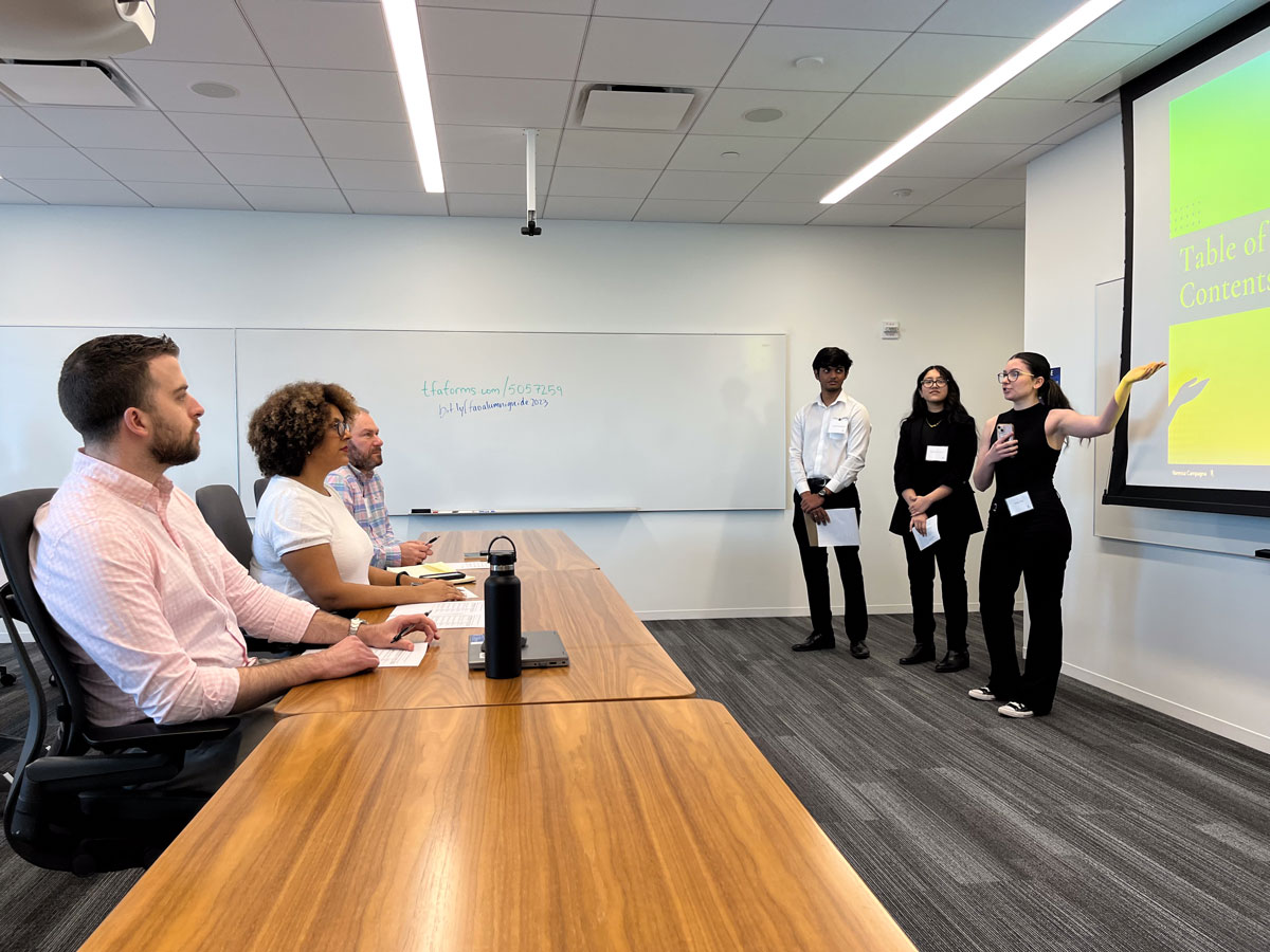 Three students stand and present in front of a projection screen while three instructors sit at a table listening